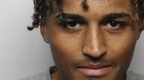 A close-up image of a police mugshot, showing a strip of the face between just above the eyebrow and just below the mouth. Daniel Heeley-Luty has ear-length hair which is twisted into tight curls. He appears to be smiling very slightly. He has some stubble on his upper lip but is mostly clean shaven, and wears an earring.