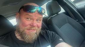 A bald man with a blonde beard has sunglasses on his forehead and is smiling at the camera while sat in a car