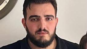 A photo of the victim, a heavy set man of around 30 with a black beard and black hair. He is wearing a black shirt and is standing in front of a cream background. 