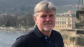 Lyndon Wright smiles into the camera. He has short grey hair and is wearing a dark polo shirt. In the background there is a river and a large ornately decorated house.