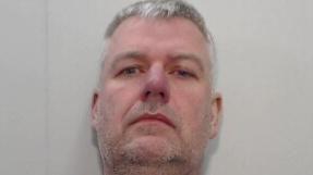 A mugshot of a middle aged man with grey hair and stubble