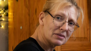 A woman in a black top wearing glasses looking at a camera. She is sitting in a room in front of a wooden door