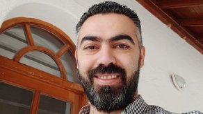 An image of Amr, who has a black beard is smiling. He is wearing a black and white checked shirt and is standing in front of a white wall with an arched wooden frame.