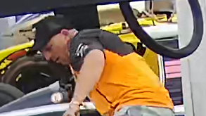 A CCTV image of a man police wish to speak to after a classic F1 car was damaged at Silverstone on Friday. The man wears a black baseball cap and orange and black T-shirt while leaning over a car.