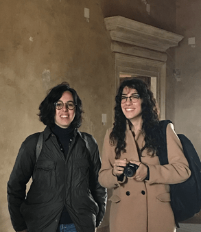 Two young women smile at the camera. One with shorter brown hair and glasses wears a dark coat and straps showing she has a rucksack on. Next to her is a longer haired brunette woman wearing a light brown coat holding a camera. They are standing in a room with beige plaster walls.
