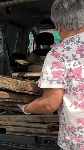 A woman in a white floral top loads some wood panels out of the back of a large car. 