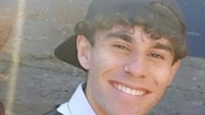 Photo of Ison Alli, who death is being investigated as murder. He has a broad smile on his face with the top row of his teeth showing. He has a black baseball cap on backwards. Mr Alli has dark curly hair. He has a nose-ring on his right nostril. He is stood in front of a wall and has a white collared shirt on.