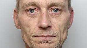A police mugshot of a man with blue eyes and grey stubble. It is cropped closely, showing just between his eyebrows and chin.