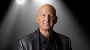 A headshot of Ian Gore, he has a shaved head and wears a glittery black jacket over a black shirt