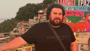 Jay Cooper pictured in a black tshirt with shoulder bag, black curly hair and beard, posing for an apparnent holiday snap