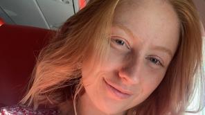 Amy has shoulder‑length, strawberry‑blonde hair styled loosely, with a few strands falling forward. Her face is angled slightly toward the camera, with light‑coloured eyes and a clear, natural complexion. The photo is very close up focusing fully on her face.