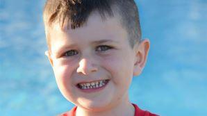 Archie smiles at the camera. He has short dark hair and is wearing a red t-shirt, with the blue waters of a swimming pool behind him.
