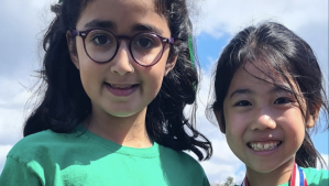 Nuria Sajjad and Selena Lau smiling outdoors against a cloudy blue sky, both wearing green T-shirts. Nuria is on the left wearing round glasses, and Selena is on the right and has a red, white, and blue medal ribbon around her neck.
