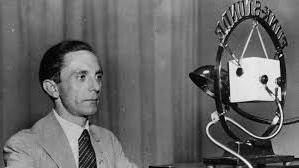 Joseph Goebbels with dark swept back hair, is sitting behind a microphone. An Anglepoise lamp is next to him. He is wearing a collar and tie with a light coloured jacket.