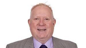 Jamie Hanlon. He is stood in front of a hite background wearing a grey suit and a pink shirt.