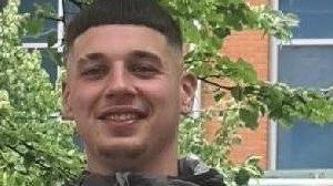 A pixelated photo of a man with cropped dark hair and wearing a grey top, smiling at the camera. There's a brick wall, a white window frame and a tree behind him.