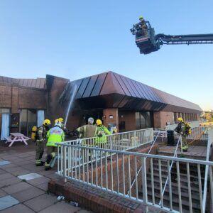 Firefighters standing on a terrace of the Bournemouth International Centre spraying water at the building's roof.