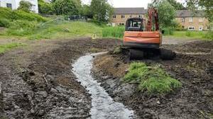 An orange digger can be seen on wet, muddy ground. Sand-coloured houses can be seen in the background.