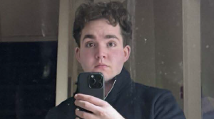 A man taking a selfie in a mirror which has marks on it. He has short wavy brown hair and is wearing a black jumper and pulling a serious expression. He is holding up a black iPhone with three camera lenses on it.