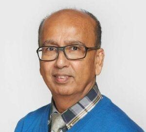 Prof Ahmed wears a black pair of glasses and a chequed shirt, grey tie and bright blue v neck jumper.  It is a professional photo and he is smiling.