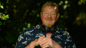A picture of Mark Mardell smiling in a garden in a floral shirt with headphones around his neck