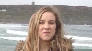 A woman with blonde hair and wearing a brown coat is standing on a beach in front of sand and rocks. The sea is in the background.
