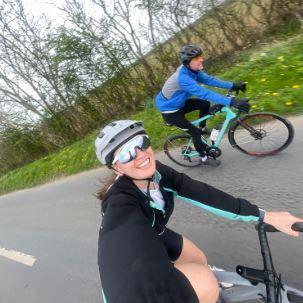 A woman cycling along a road and taking a selfie. She is wearing a helmet and silver cycle glasses and a black and blue jacket. Next to her is a man cycling alongside her wearing a helmet and blue coat.