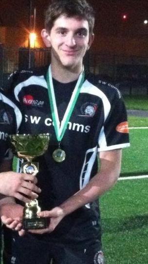 Young man in dark blue rugby outfit with sw comms logo on the front holding a cup and standing on a rugby pitch.