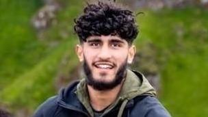 Mohammed Afzal, who has a dark beard and curly hair, smiles while climbing a hill. He is wearing a dark jacket with a green hood.