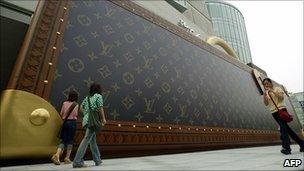 Shoppers walk beside a giant Louis Vuitton advertisement shaped like a briefcase