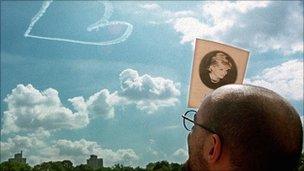 A man attending a memorial service for Diana, Princess of Wales in New York uses the program to shield his eyes from the sun as he watches a sky-writing aircraft sketch a heart in the sky