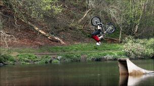 A mountain biker flips in the air froma ramp into the river.