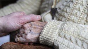 A close-up picture of a carer reaching out to hold the hand of an elderly person in a cardigan