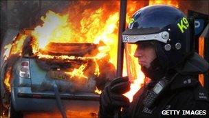 Police officer stands in front of a car on fire in Hackney