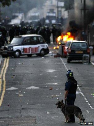 A police dog handler stands behind officers in riot gear blocking a road near a burning car on a street in Hackney