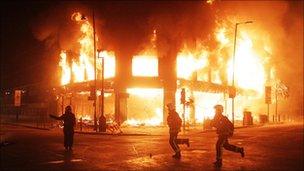 Fire fighters and riot police silhouetted against a burning building as they run past it, Tottenham - Pic: PA