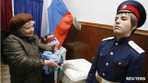 A woman carrying a child casts her ballot next to a Russian cossak (R) at a polling station in Rostov-on-Don