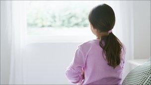 Young female child (girl, youth) sitting alone on the edge of a bed and looking out through a bedroom window