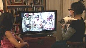 Two French people sit in front of a TV showing archive footage of the Queen