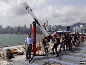 A man poses for a photograph as people queue up along the Hong Kong harbour waterfront to look through a telescope