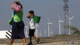 A woman and a boy walk past wind turbines and an electricity tower in La Ventosa, Mexico