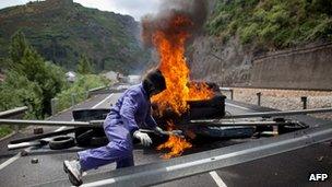 A masked miner climbs over a barricade which includes a burning tyre