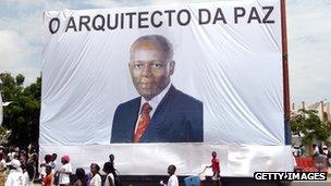 People walk in front of a large portrait of Angola President Jose Eduardo de Santos with text reading 'The Architect of Peace', in the streets of Luanda