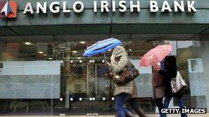 Pedestrians holding umbrellas pass in front of an Anglo Irish Bank branch