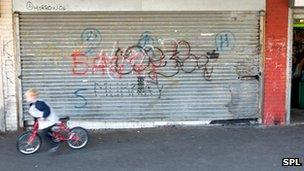 Boy on bike rides past boarded-up shop