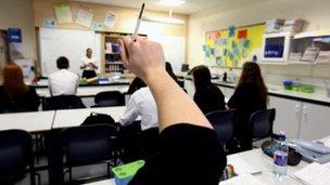 Schoolchildren raise their hands in a classroom as a teacher stands by a whiteboard