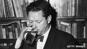 Dylan Thomas drinking a glass of beer and smoking while seated at a desk with stacks of his books of poetry, New York City, c. 1950