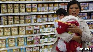 File photo: a woman holding a baby stands in front of a shelf displaying milk powder products at a supermarket in Beijing, 20 May 2013