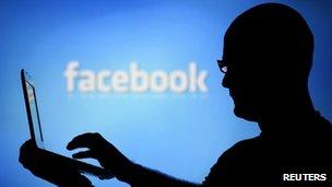 A man with a laptop is silhouetted against a screen with a Facebook logo in the background 14 August 2013