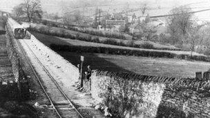 Two children sit on a wall waiting for a train in this black-and-white postcard from the 1930s.